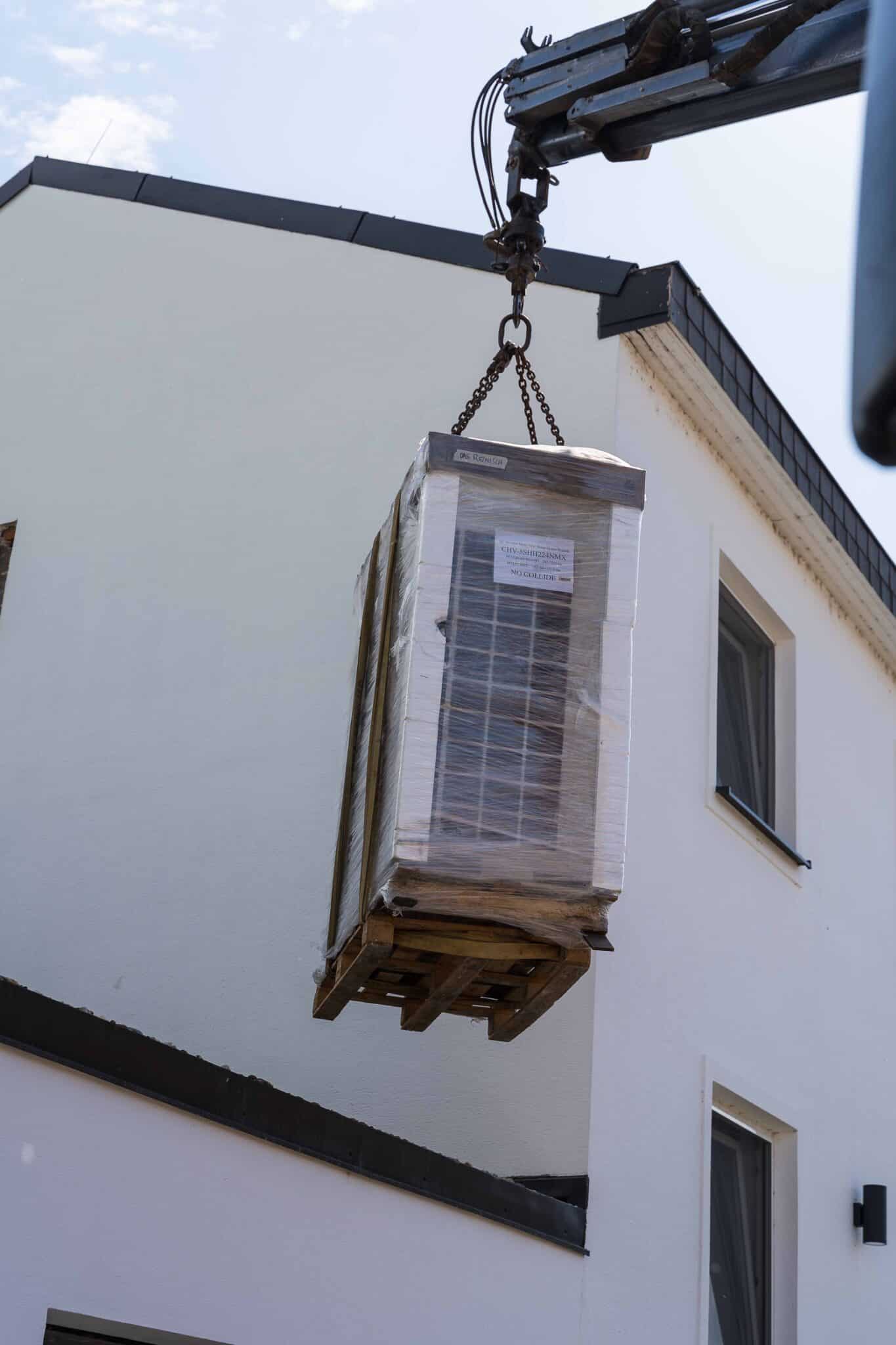 Eine große Klimaanlagen-Außeneinheit wird mit einem Kran in Folie verpackt auf ein Dach gehoben.