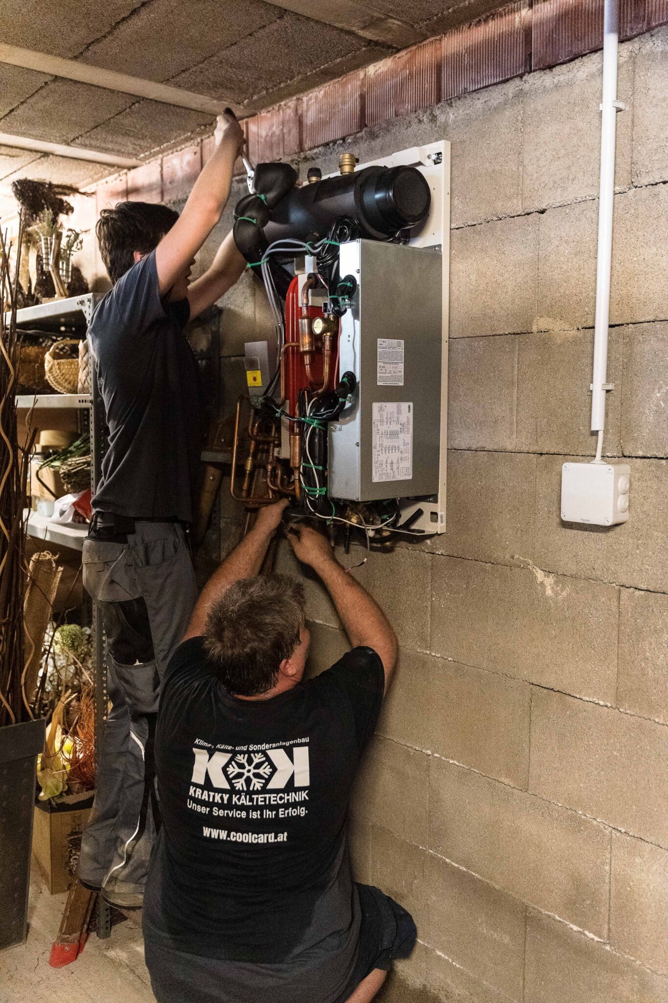 Zwei Handwerker montieren eine Hydro-Box an einer Betonwand, das Gehäuse ist geöffnet und die Leitungen werden angeschlossen.